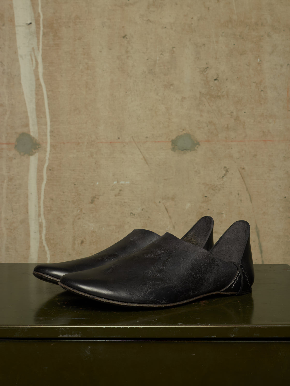 A pair of black leather slip-on shoes with pointed toes, displayed on a dark reflective surface against a textured beige wall.