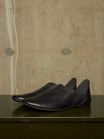 A pair of black leather slip-on shoes with pointed toes, displayed on a dark reflective surface against a textured beige wall.