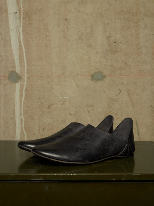 A pair of black leather slip-on shoes with pointed toes, displayed on a dark reflective surface against a textured beige wall.