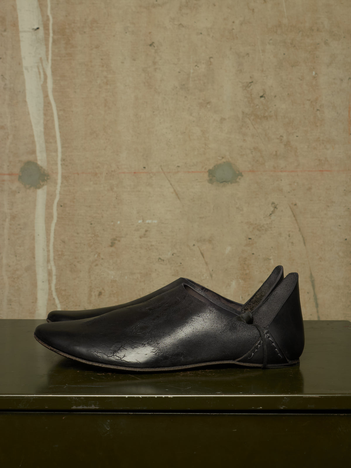 A pair of black leather slip-on shoes with a pointed toe and heel tab, displayed on a dark green surface against a textured beige concrete wall.