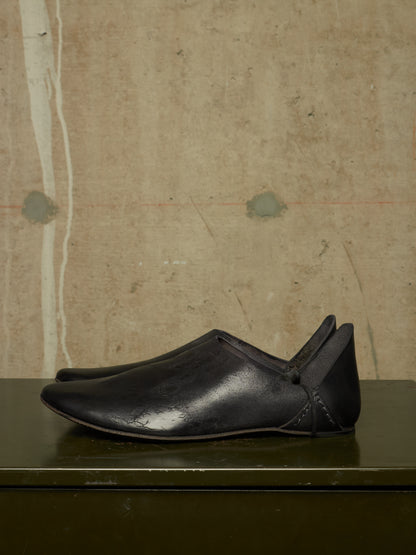 A pair of black leather slip-on shoes with a pointed toe and heel tab, displayed on a dark green surface against a textured beige concrete wall.