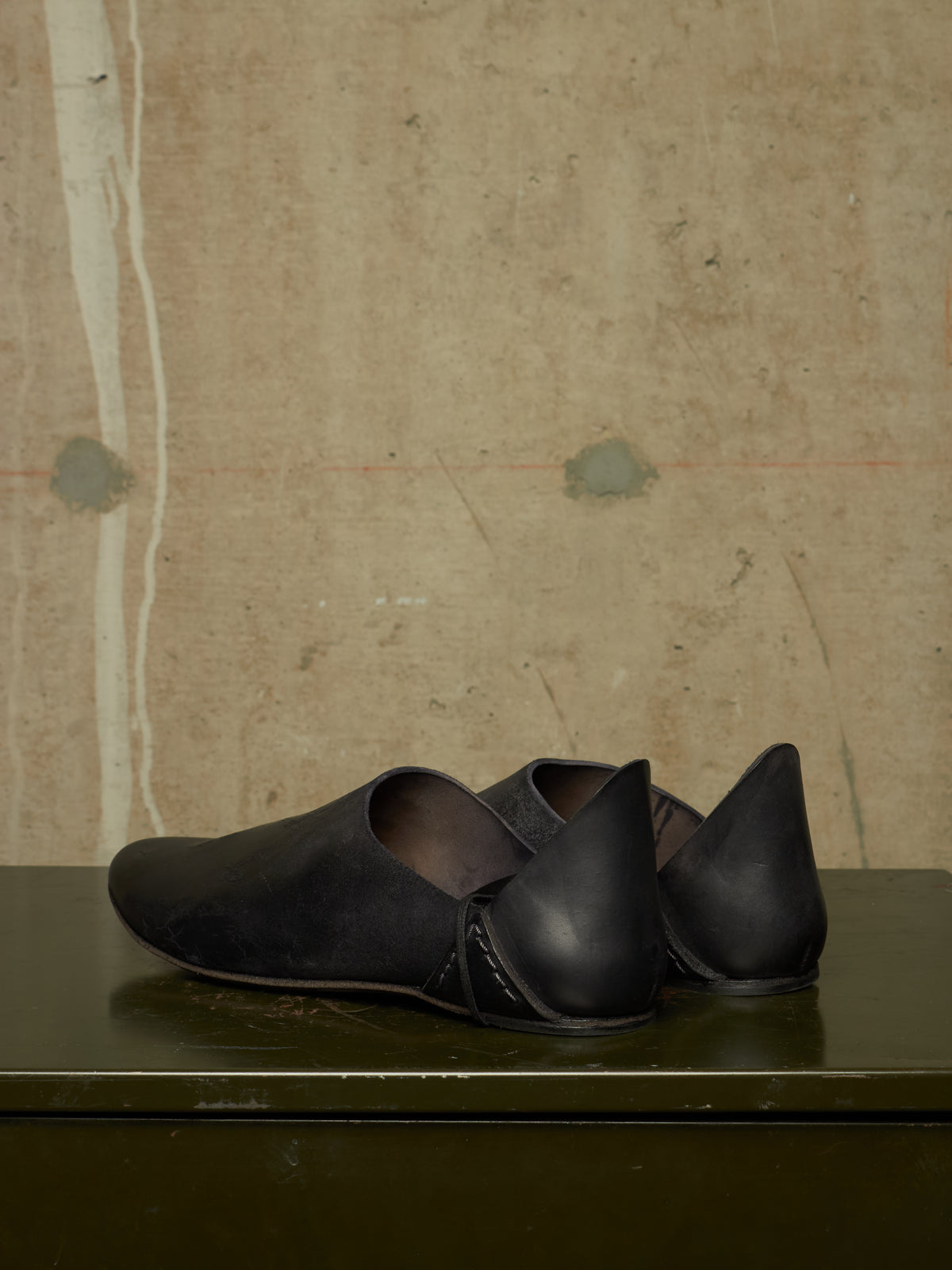 A pair of black leather slip-on shoes with pointed heels, displayed on a dark green surface against a textured beige concrete wall.