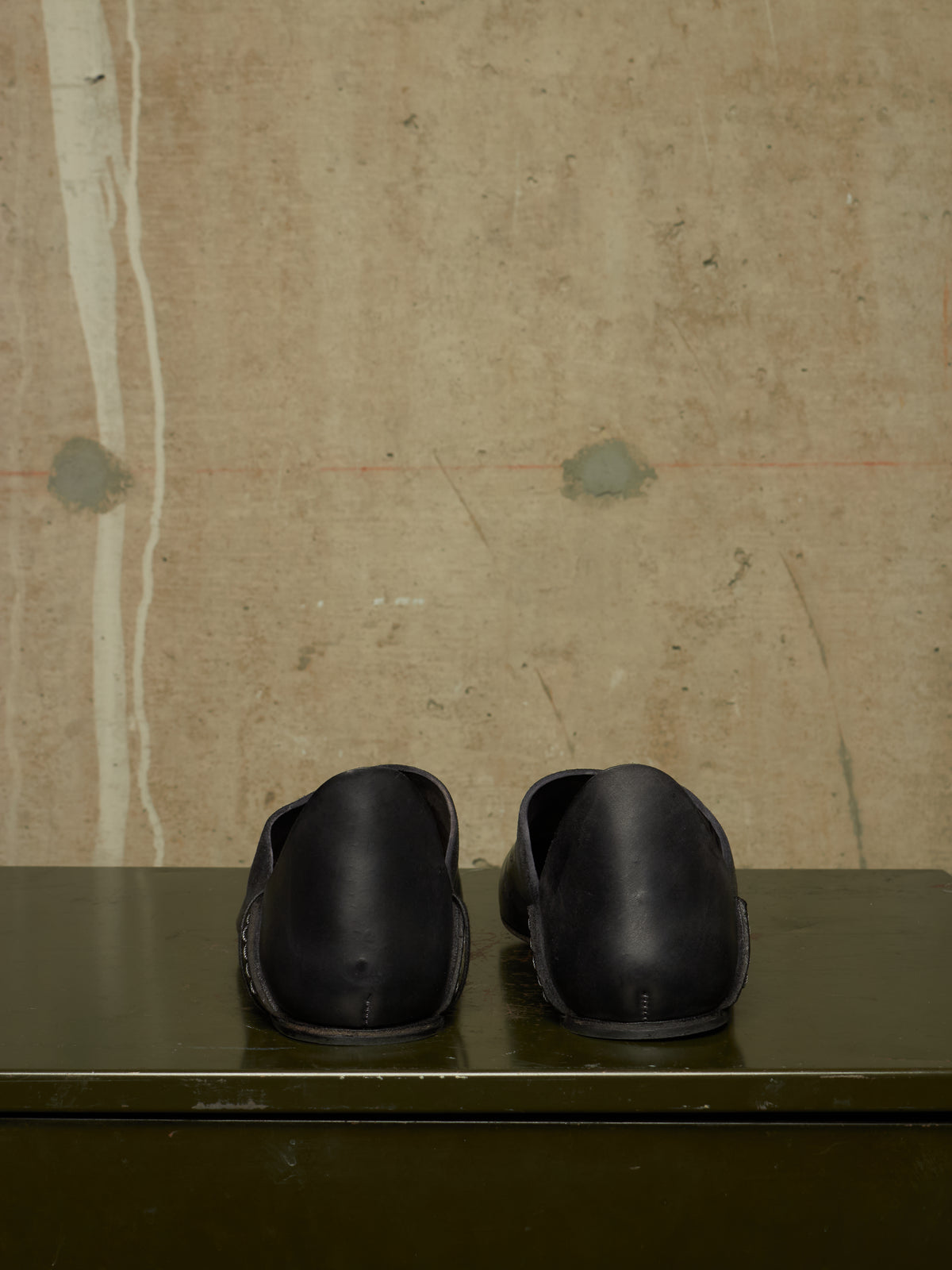 A pair of black leather slip-on clogs shown from the back, resting on a dark green wooden surface against a textured beige concrete wall.