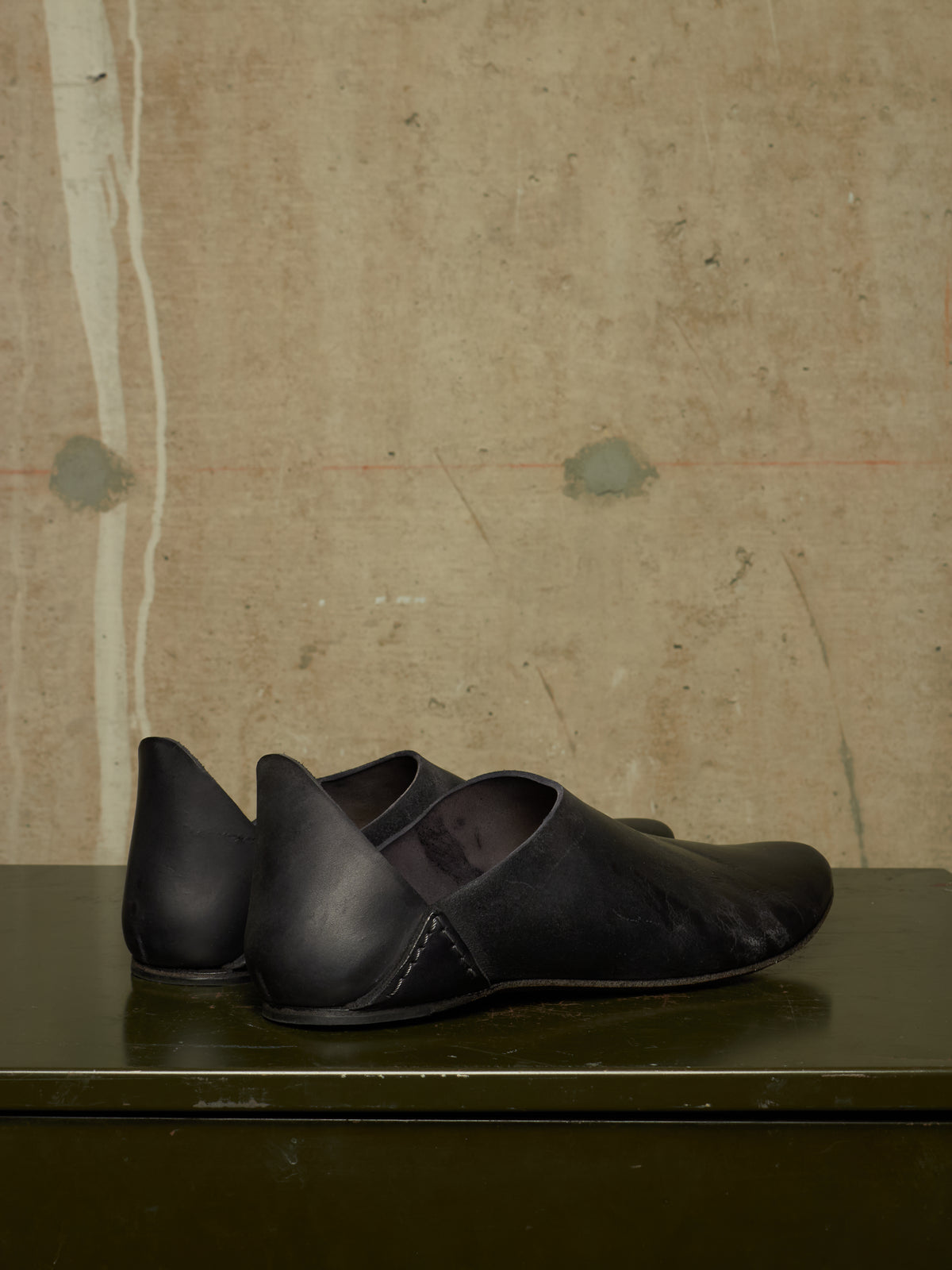 A pair of black leather slip-on shoes with a pointed toe and contrast stitching, resting on a dark green surface against a textured beige wall.