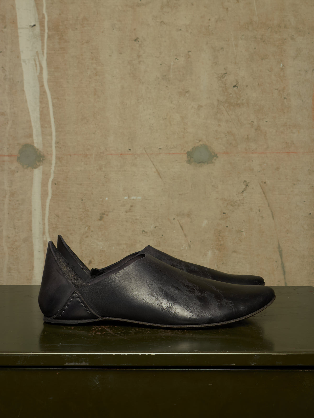 A pair of black leather slip-on shoes with pointed toes, displayed on a dark green surface against a textured beige wall.