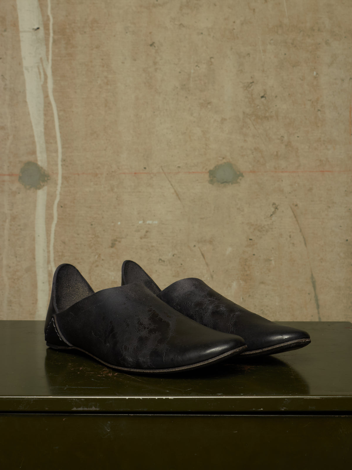 A pair of black leather slip-on shoes with pointed toes, displayed on a dark green surface against a textured beige concrete wall.