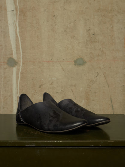 A pair of black leather slip-on shoes with pointed toes, displayed on a dark green surface against a textured beige concrete wall.