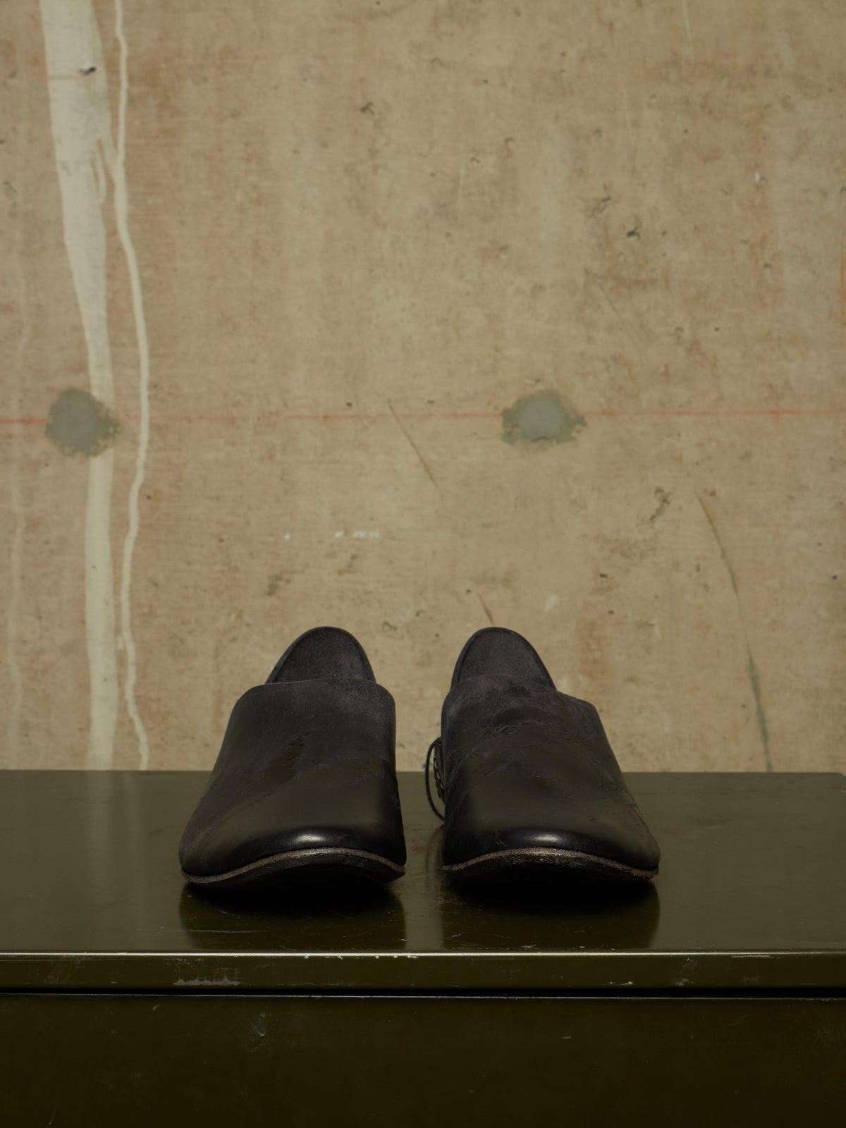 A pair of black leather slip-on shoes with a matte finish, displayed on a dark green surface against a textured beige concrete wall.