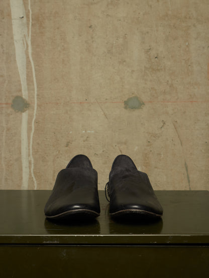 A pair of black leather slip-on shoes with a matte finish, displayed on a dark green surface against a textured beige concrete wall.