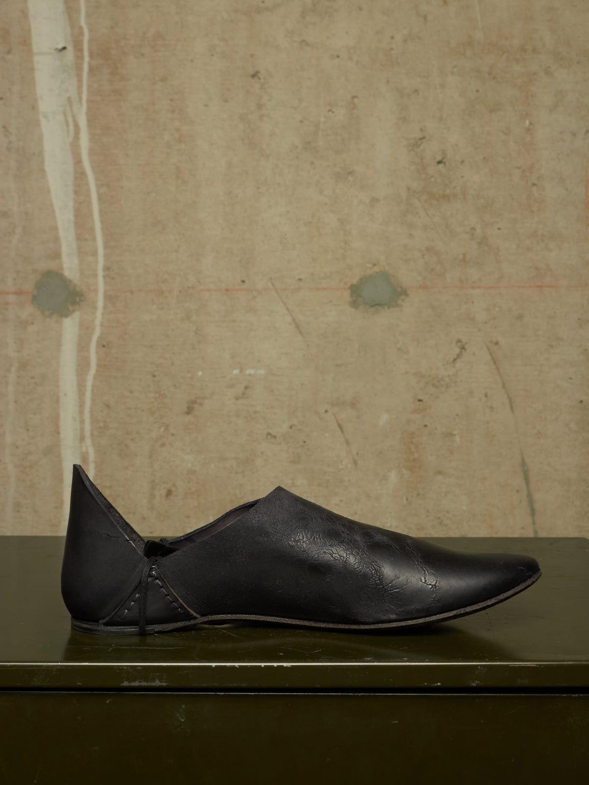 A single black leather slip-on shoe with a pointed toe and decorative stitching, displayed on a dark green surface against a textured beige wall.