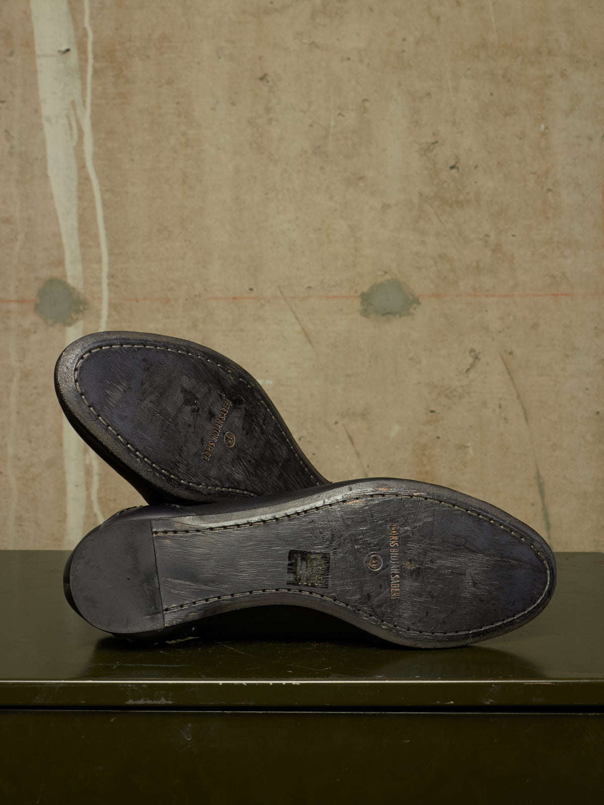 A pair of black leather shoe soles shown from above, resting on a dark green surface against a textured beige wall. The brand name 'Burberrys' is faintly visible on the soles.