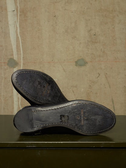 A pair of black leather shoe soles shown from above, resting on a dark green surface against a textured beige wall. The brand name 'Burberrys' is faintly visible on the soles.