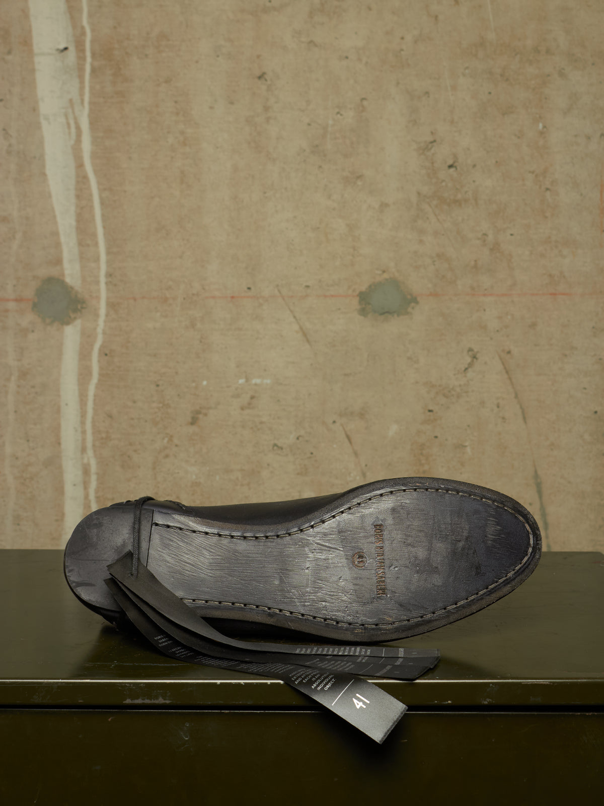 A single dark leather thong sandal shown from the underside, resting on a dark green surface against a textured beige wall background.