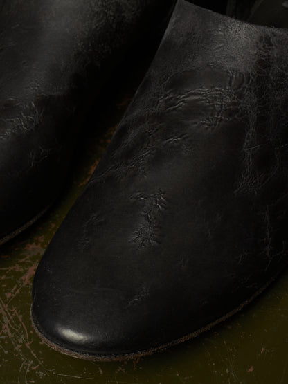 A close-up of a black leather boot with a distressed, cracked texture, resting on a dark olive green surface.