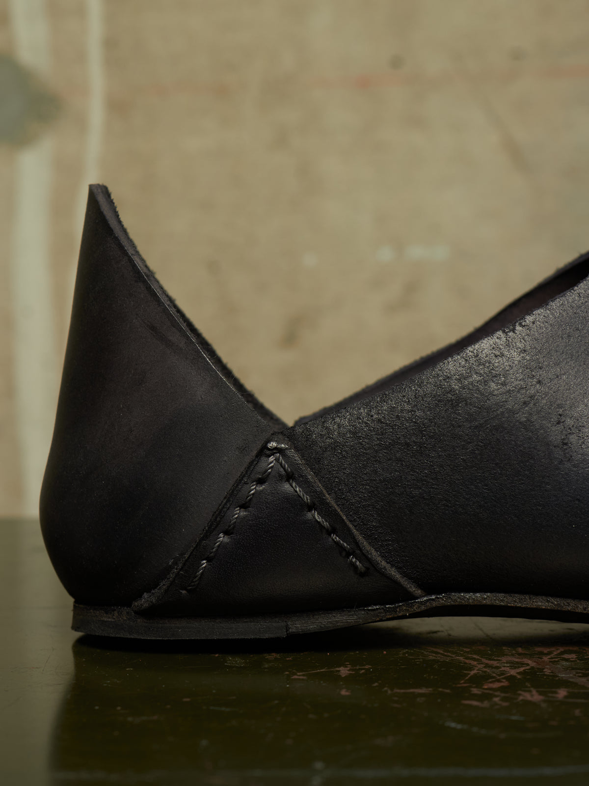 A close-up of a black leather slip-on shoe with a pointed toe and visible hand-stitched detailing, resting on a dark reflective surface against a textured beige background.