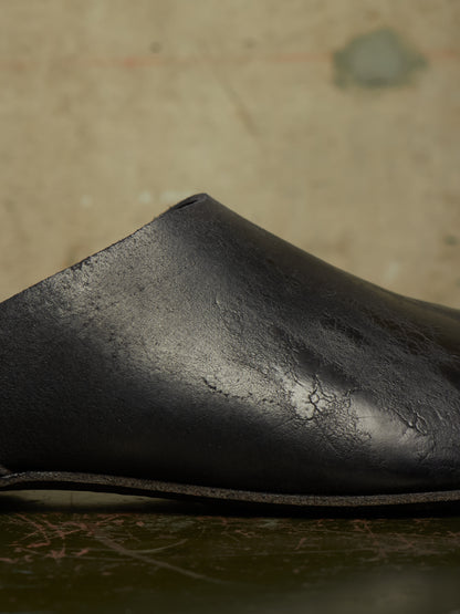 A close-up of a black leather shoe or boot showing the textured upper and sole edge, photographed against a warm beige background.