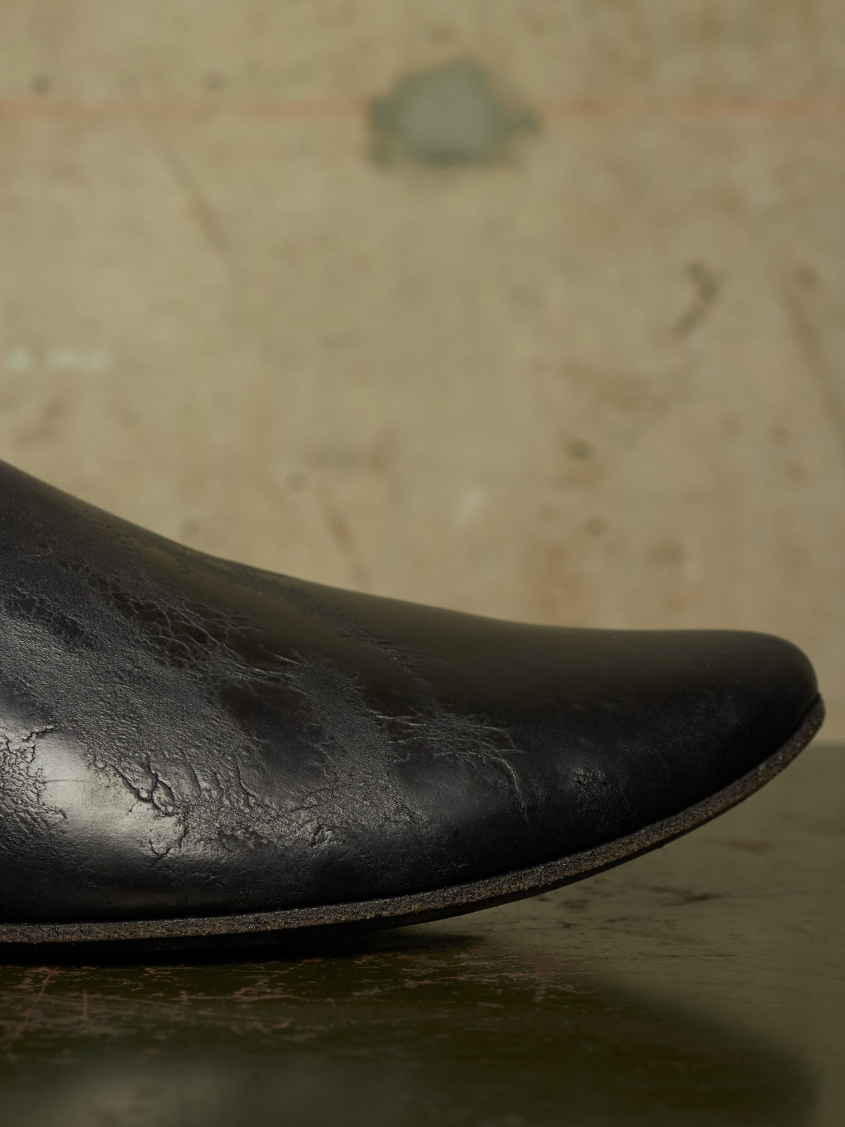 Close-up of a black leather boot toe showing textured, distressed leather and a dark sole against a warm, out-of-focus beige background.