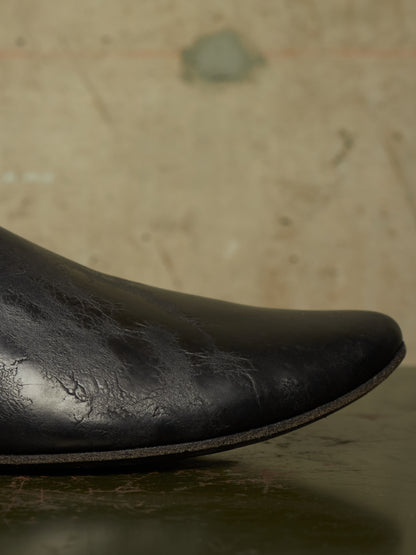 Close-up of a black leather boot toe showing textured, distressed leather and a dark sole against a warm, out-of-focus beige background.