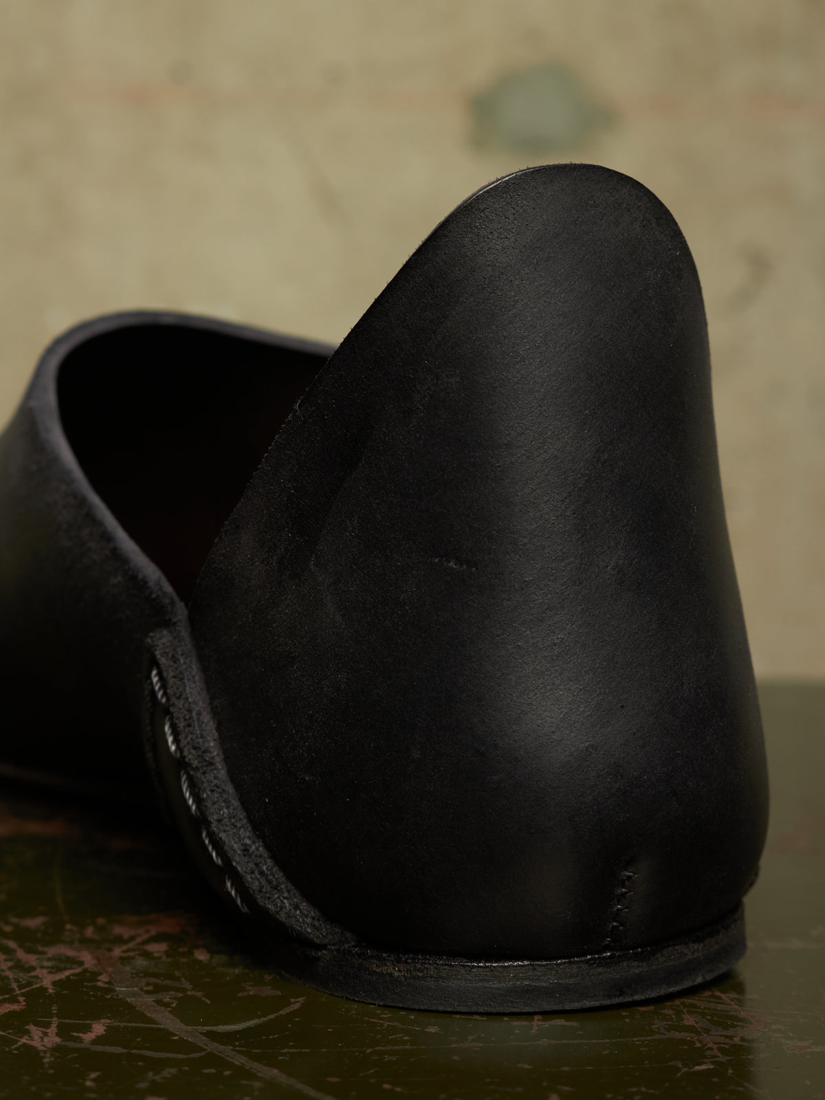 A close-up shot of a black leather slip-on shoe, showing the back of the footbed and the sole, resting on a dark textured surface against a neutral background.