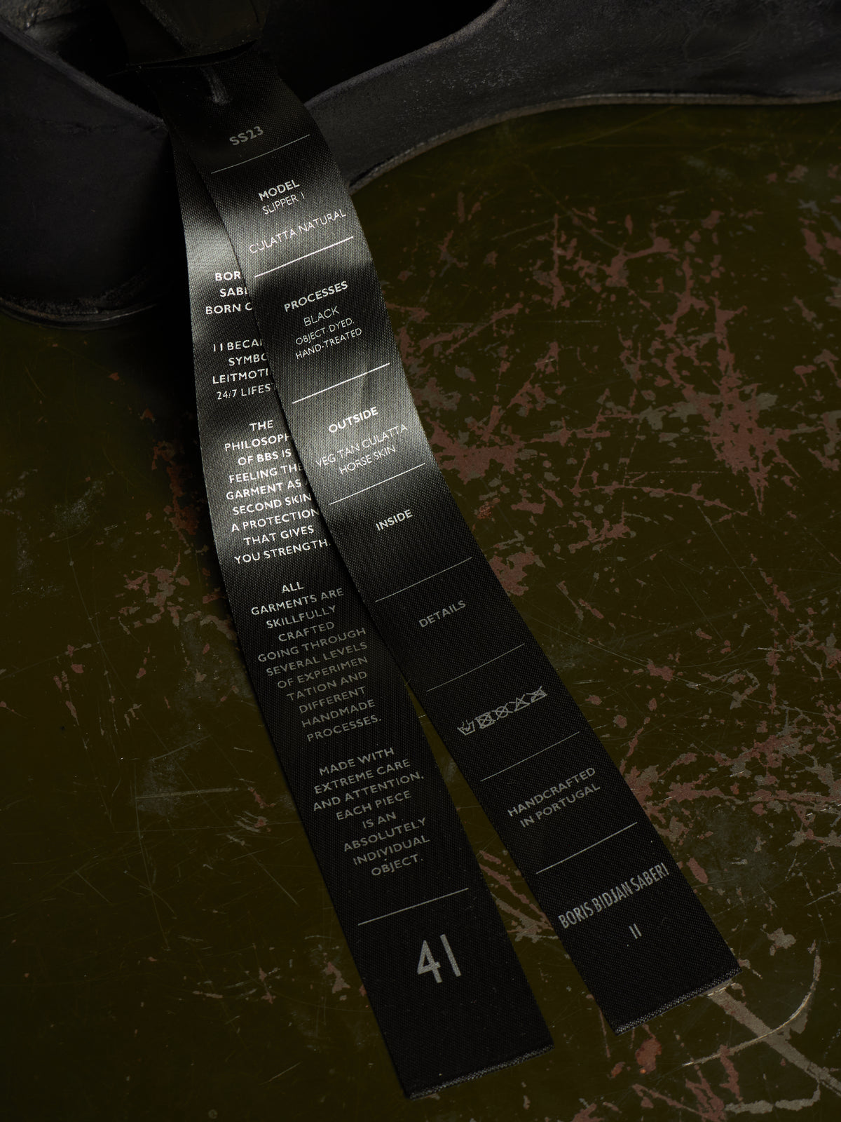 A close-up of a black fabric tag printed with white text, laid over a distressed olive green surface.