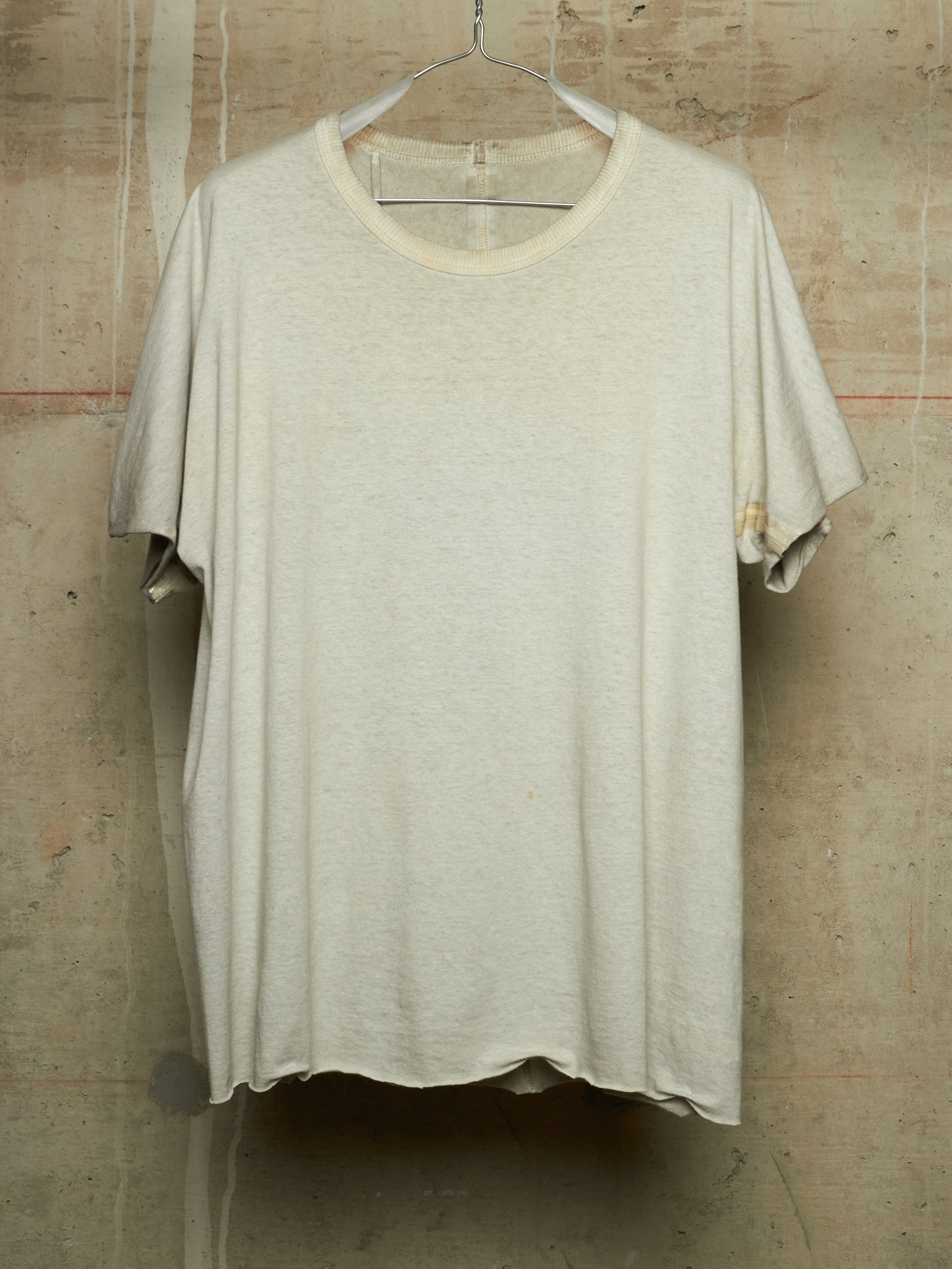 A light beige oversized short-sleeve t-shirt with a raw, distressed hem and sleeve edges, hanging on a wire hanger against a textured concrete wall.