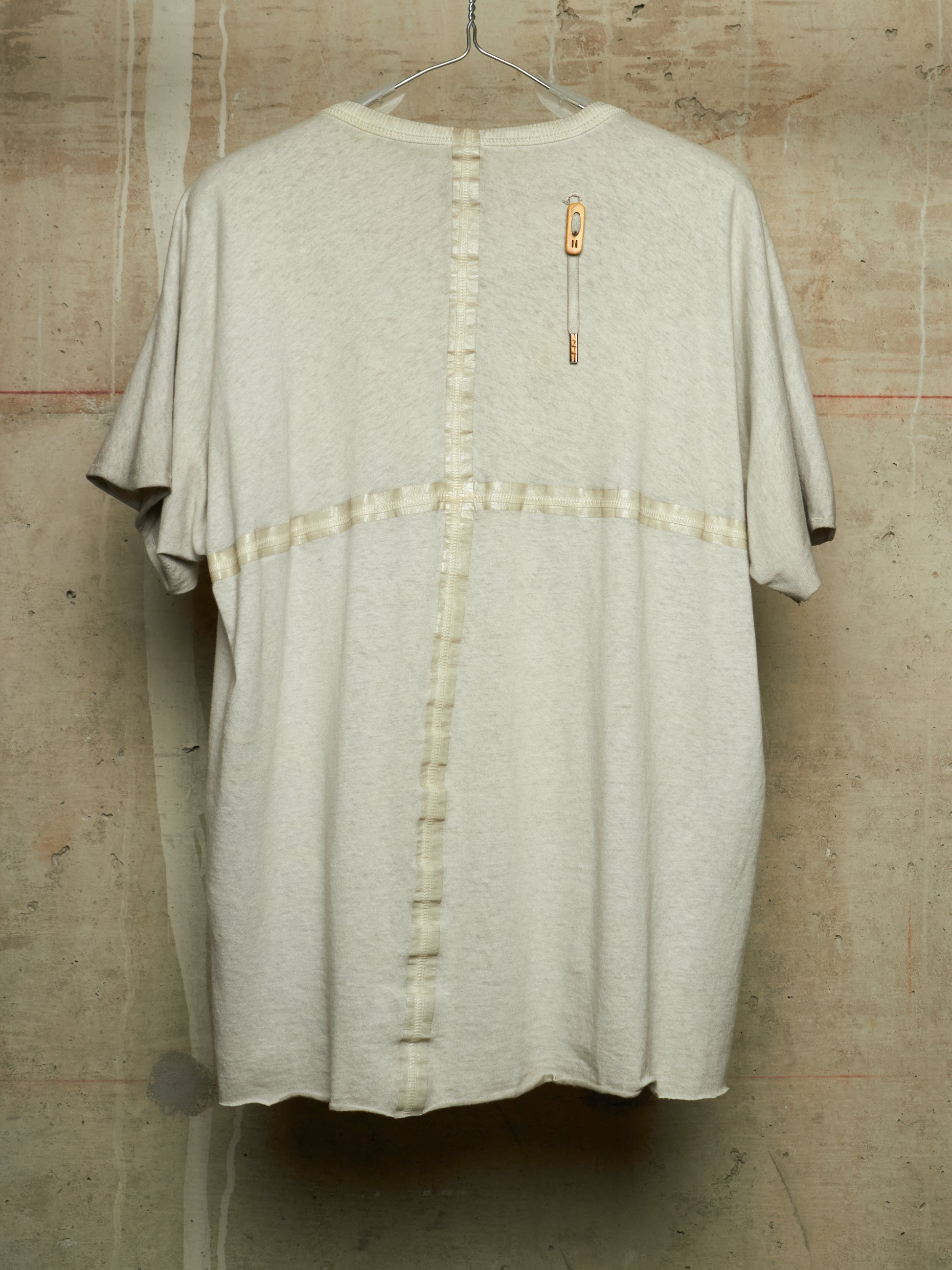A light beige oversized short-sleeve t-shirt with a vertical button placket and a horizontal button band across the chest, hanging against a textured concrete wall.