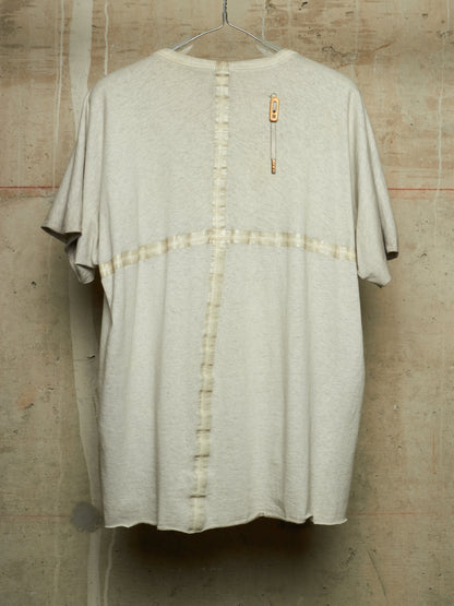 A light beige oversized short-sleeve t-shirt with a vertical button placket and a horizontal button band across the chest, hanging against a textured concrete wall.