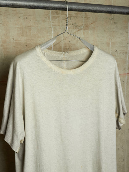 A cream-colored short-sleeve crewneck t-shirt hanging on a clear plastic hanger from a dark metal rod, set against a textured concrete wall.