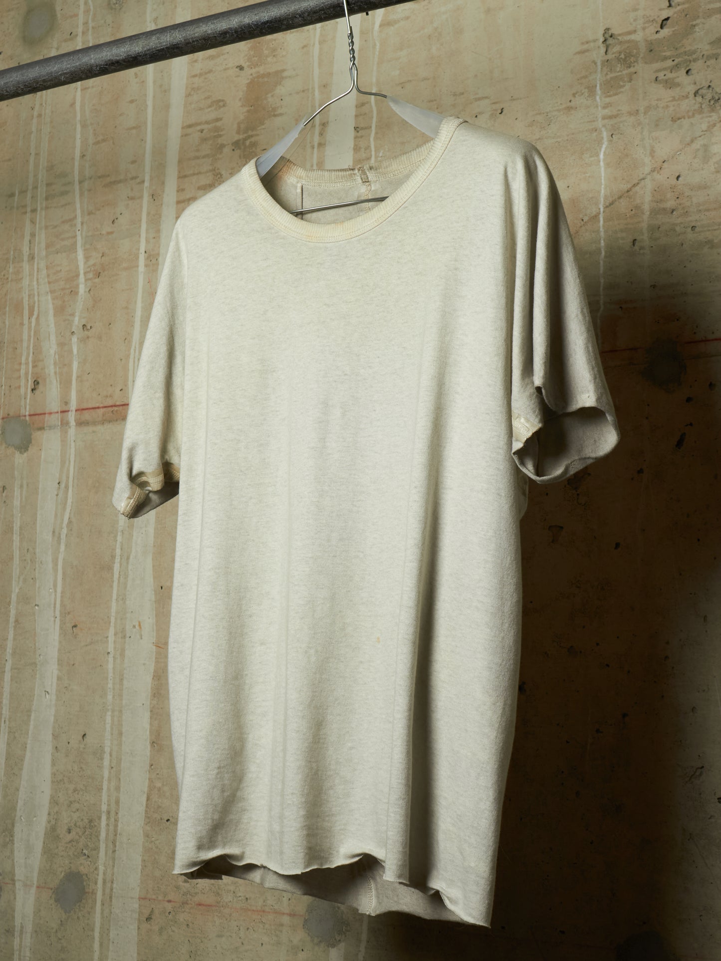 A light beige short-sleeve crewneck t-shirt hanging on a metal hanger from a dark industrial rod, set against a textured concrete wall.