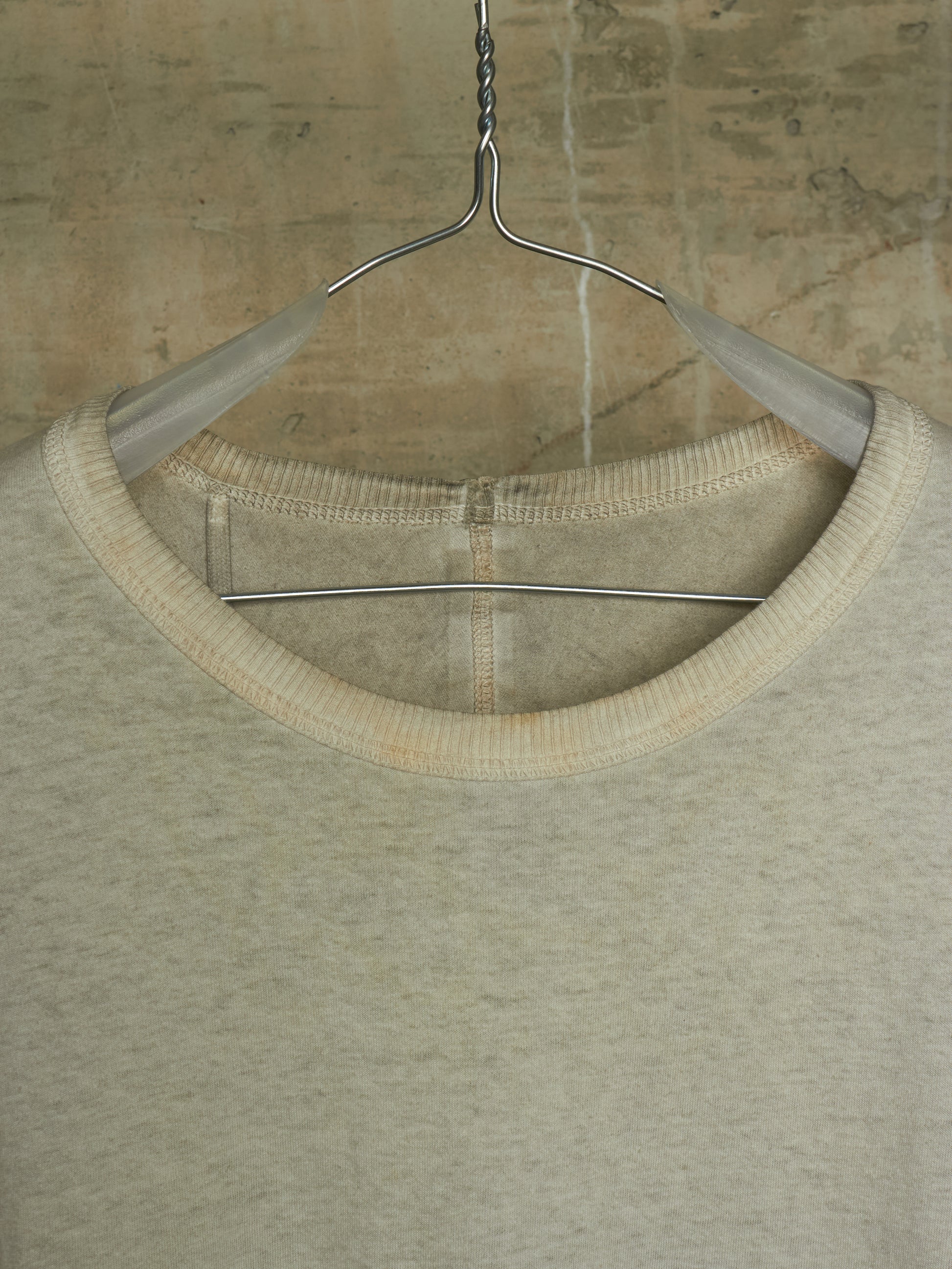 A close-up of a light beige crewneck knit top hanging on a metal hanger against a textured concrete wall.