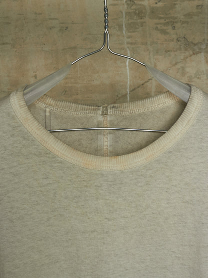 A close-up of a light beige crewneck knit top hanging on a metal hanger against a textured concrete wall.