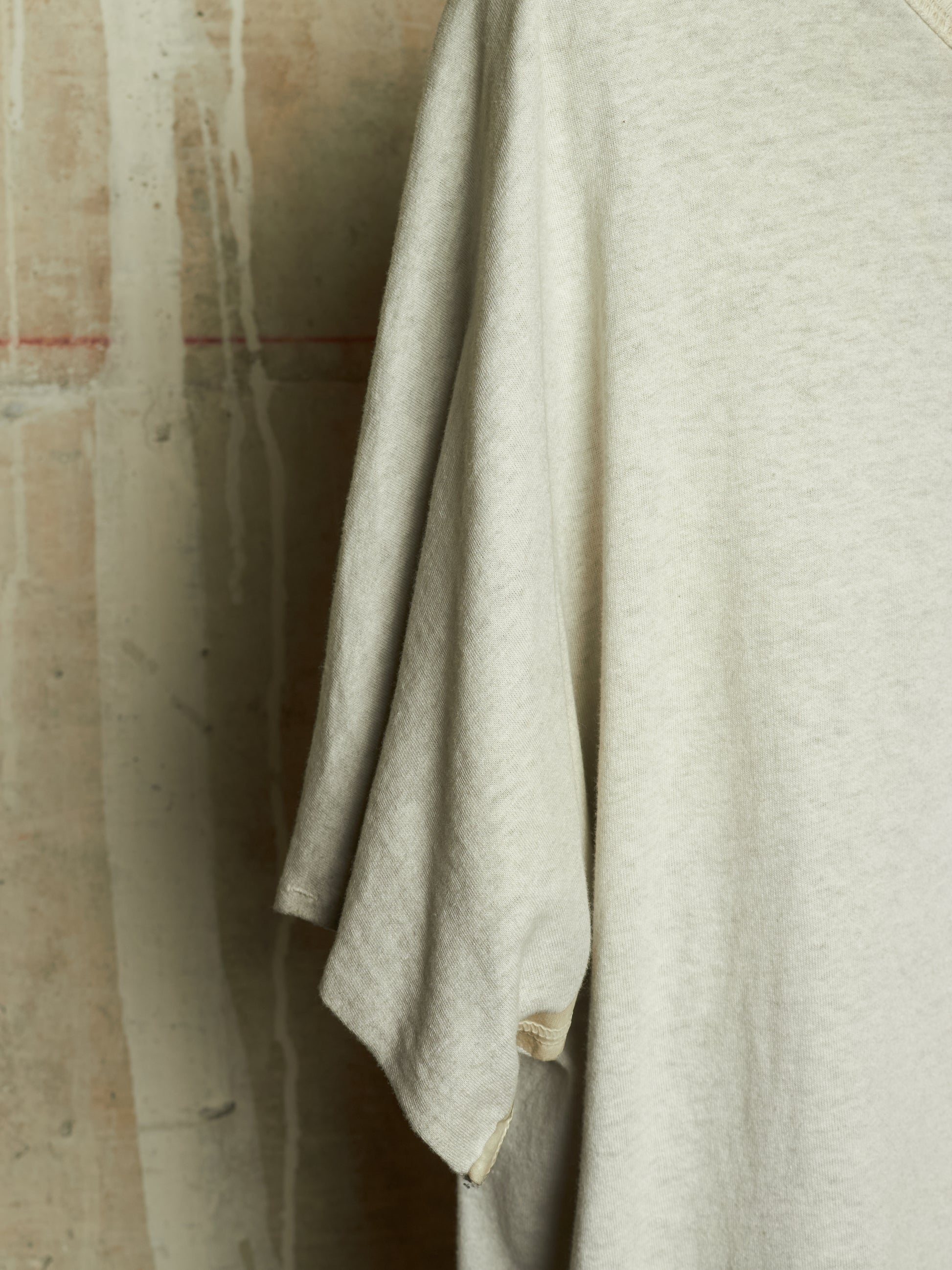 A close-up of a light heather gray or off-white t-shirt hanging against a textured concrete wall.