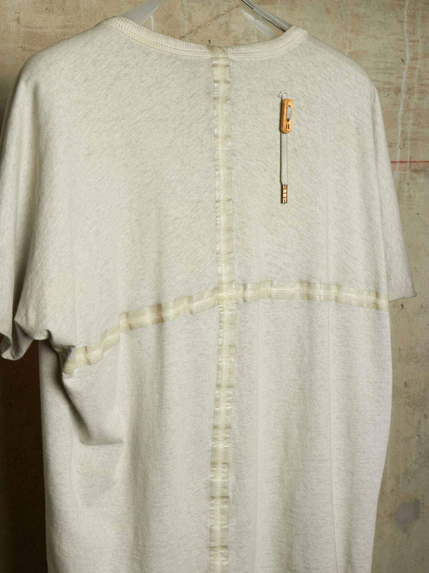 A cream-colored short-sleeve knit top hanging on a hanger against a textured concrete wall. The back of the shirt features a vertical seam with button-like stitching down the center, a horizontal seam crossing the chest, and a small orange-and-white safety pin detail on the upper back.