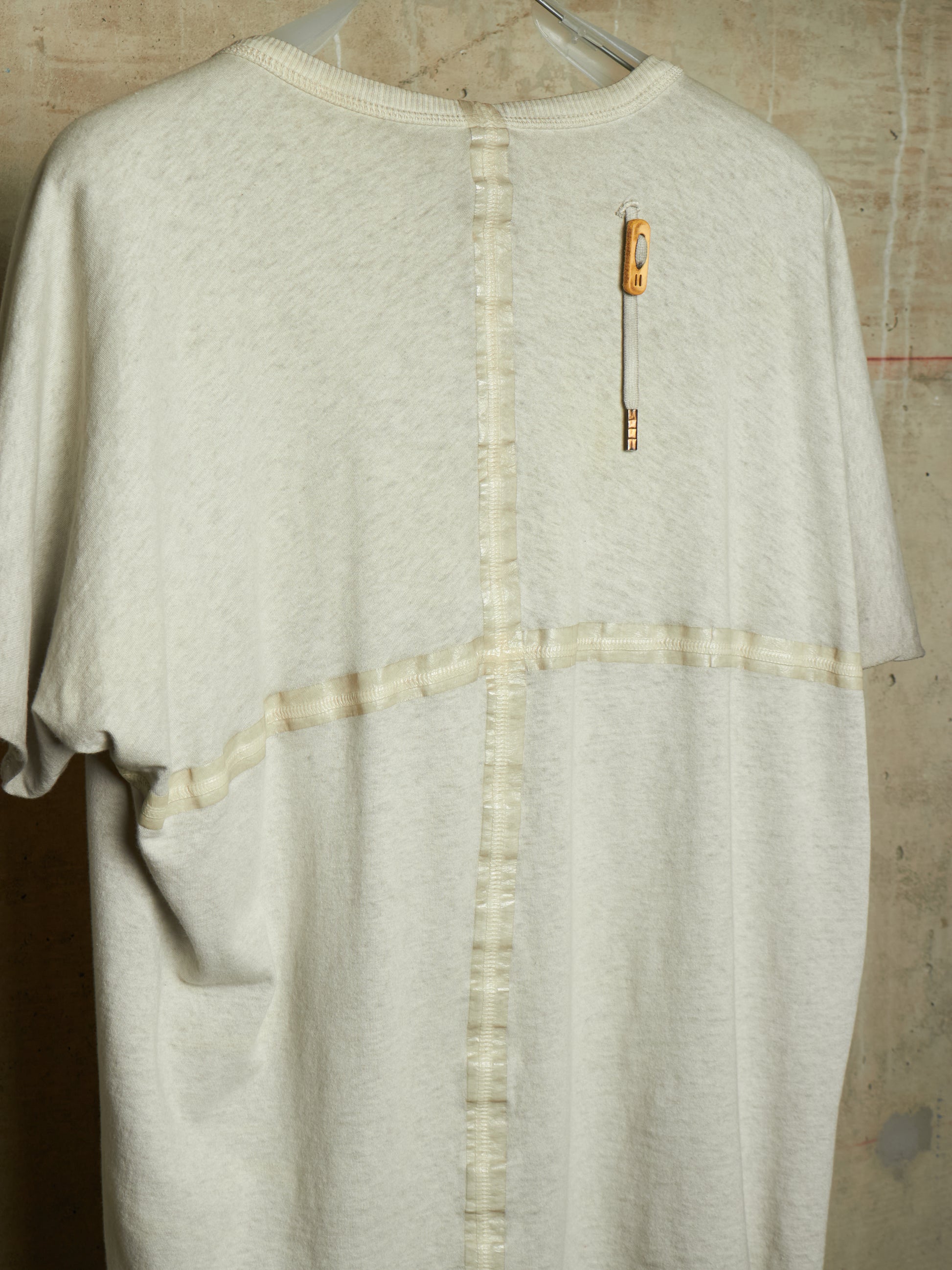 A cream-colored short-sleeve knit top hanging on a hanger against a textured concrete wall. The back of the shirt features a vertical seam with button-like stitching down the center, a horizontal seam crossing the chest, and a small orange-and-white safety pin detail on the upper back.