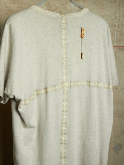 A cream-colored short-sleeve knit top hanging on a hanger against a textured concrete wall. The back of the shirt features a vertical seam with button-like stitching down the center, a horizontal seam crossing the chest, and a small orange-and-white safety pin detail on the upper back.