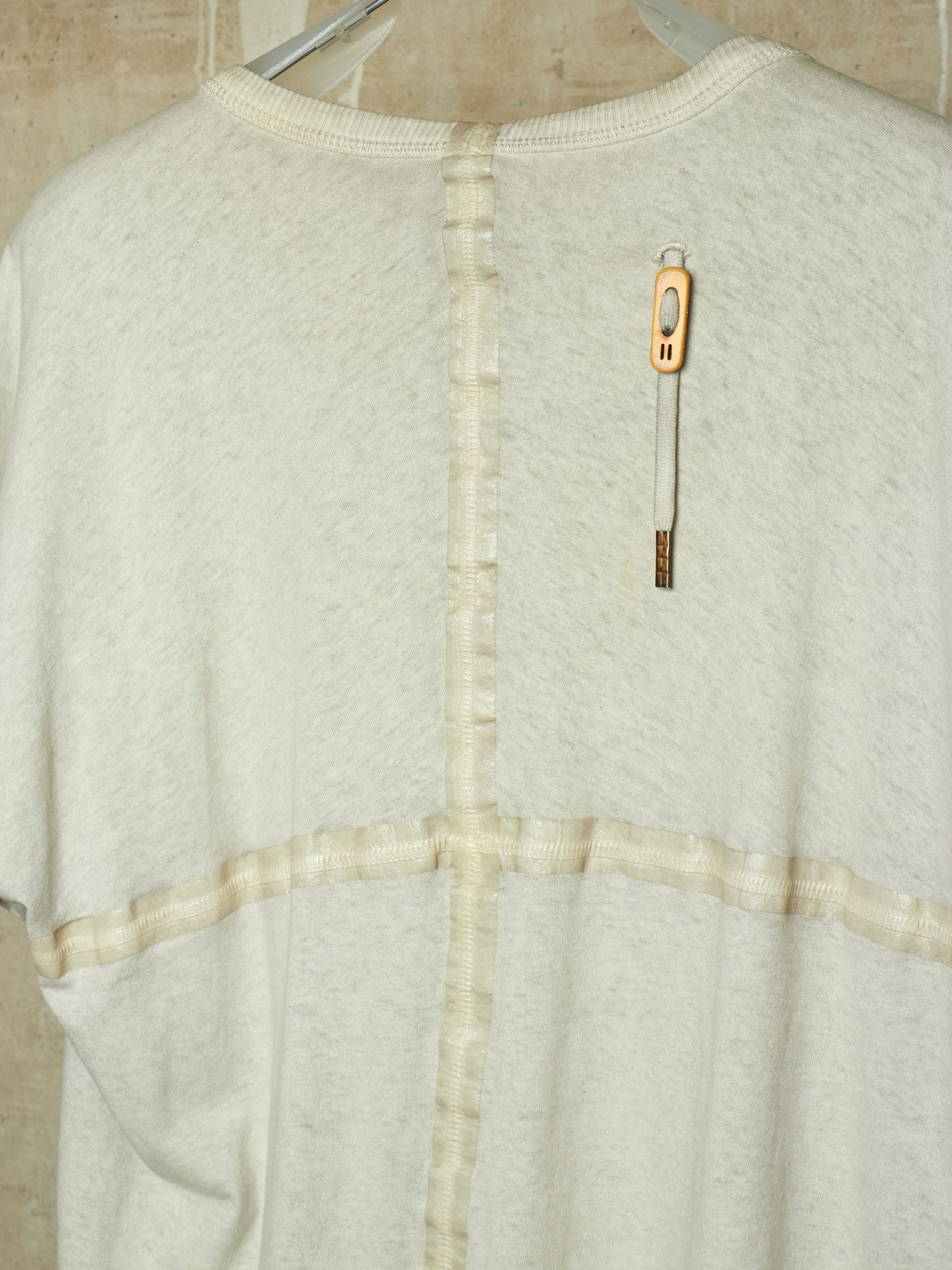 A close-up of the back of a cream-colored knit sweater with a unique vertical seam detail and a decorative gold-toned tag attached near the upper back.