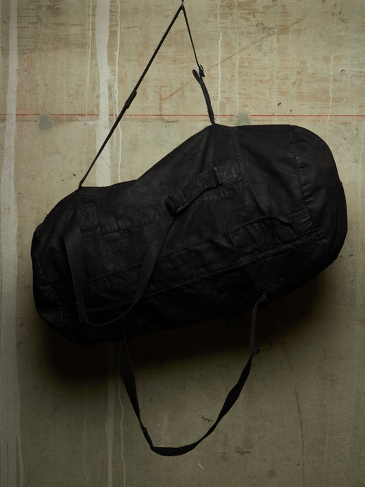 A black cylindrical duffel bag with a long shoulder strap, hanging against a textured concrete wall.