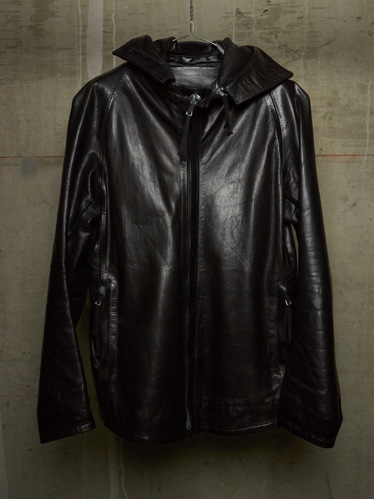 A black leather hooded jacket with a front zipper, hanging on a hanger against a textured concrete wall.