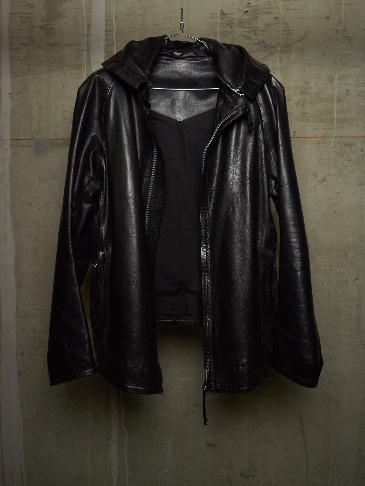 A black leather hooded jacket with a front zipper, hanging against a textured concrete wall.