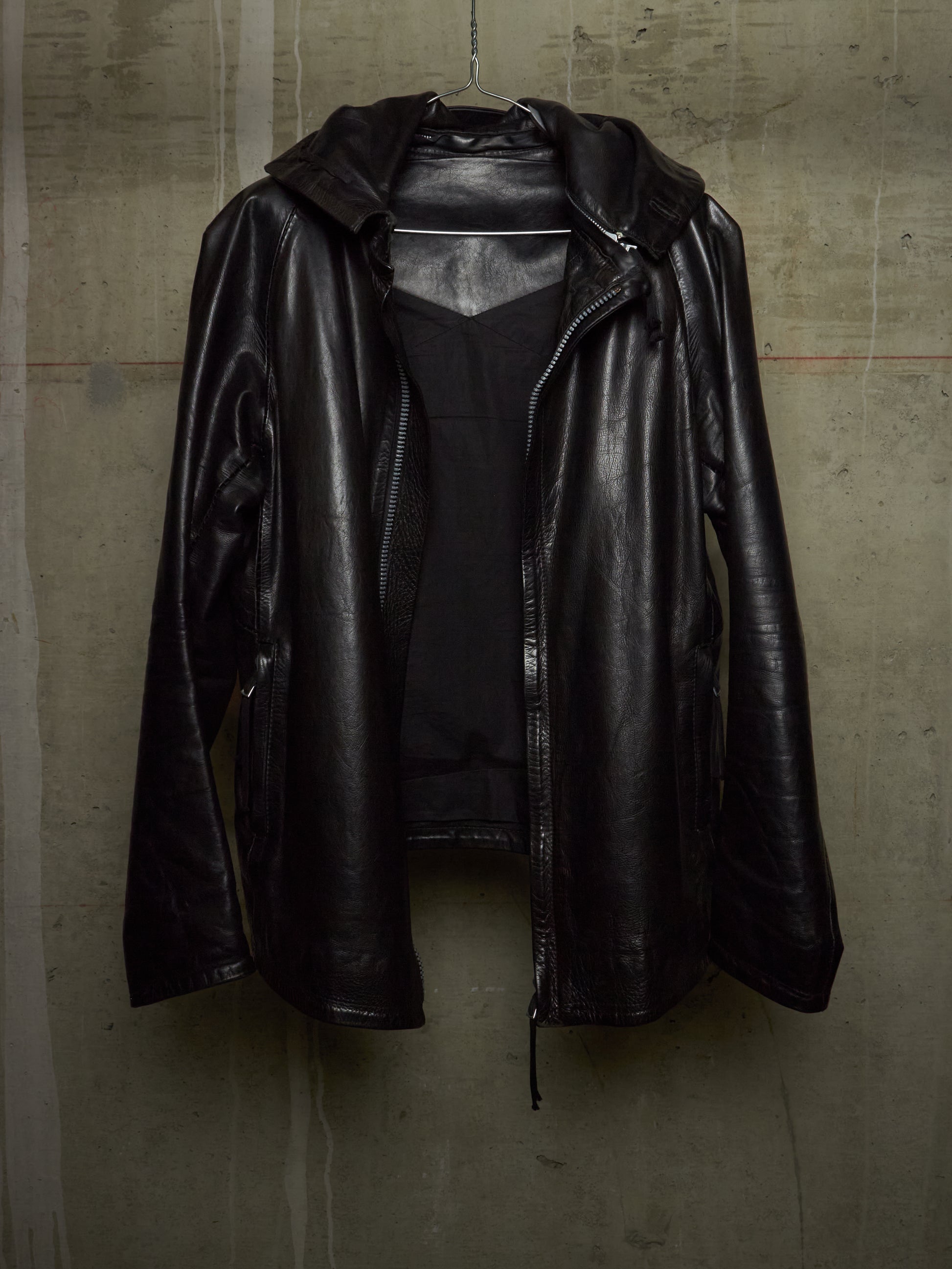 A black leather hooded jacket with a front zipper, hanging against a textured concrete wall.