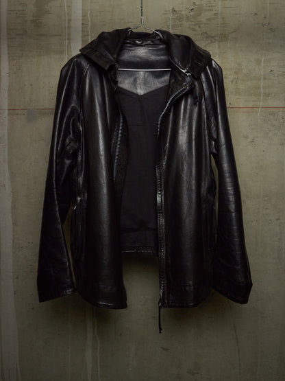 A black leather hooded jacket with a front zipper, hanging against a textured concrete wall.
