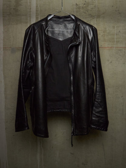 A black leather jacket with a front zipper, hanging on a wire hanger against a textured concrete wall.