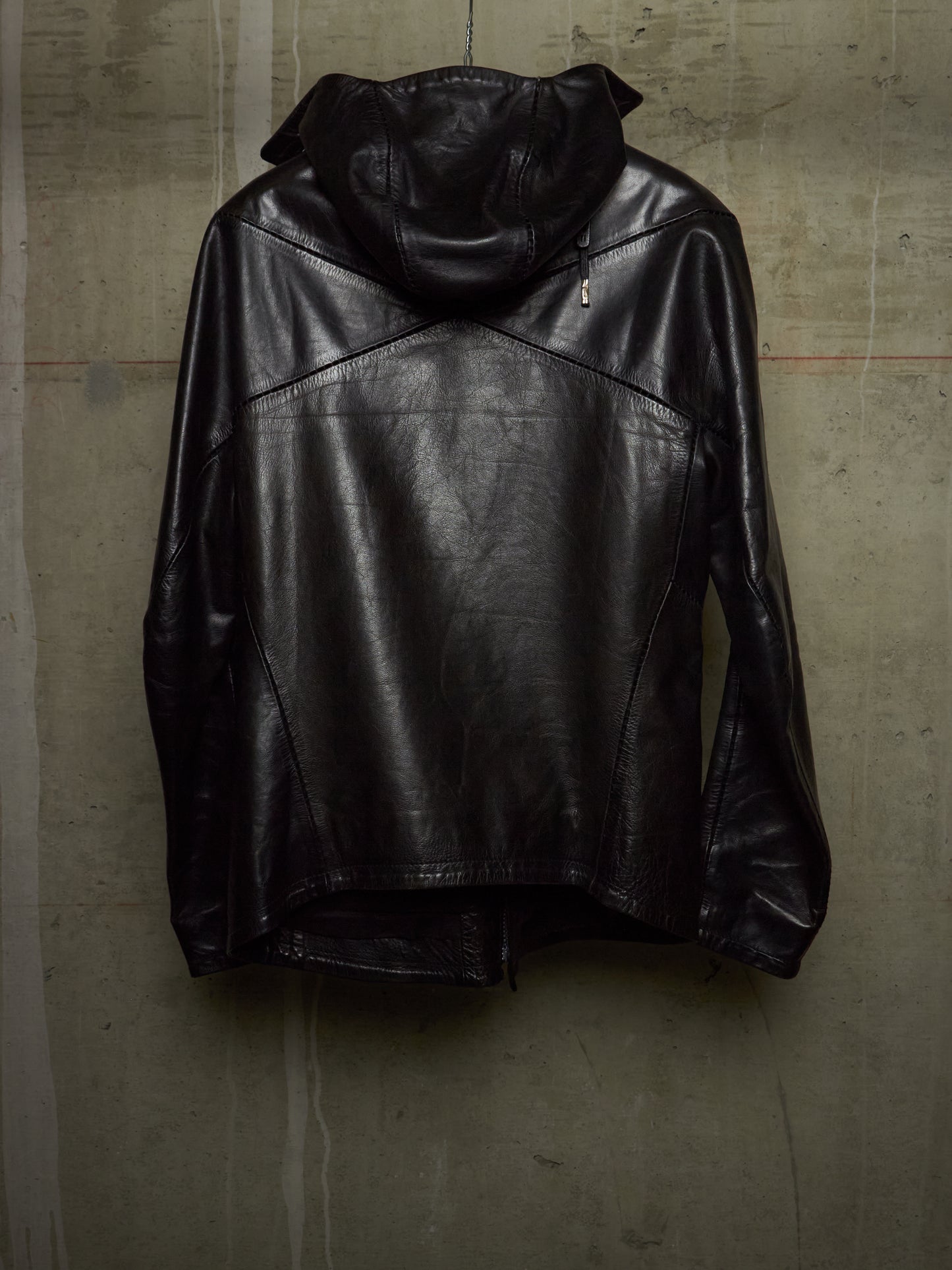 A black leather hooded jacket with diagonal seam details, hanging against a textured concrete wall.