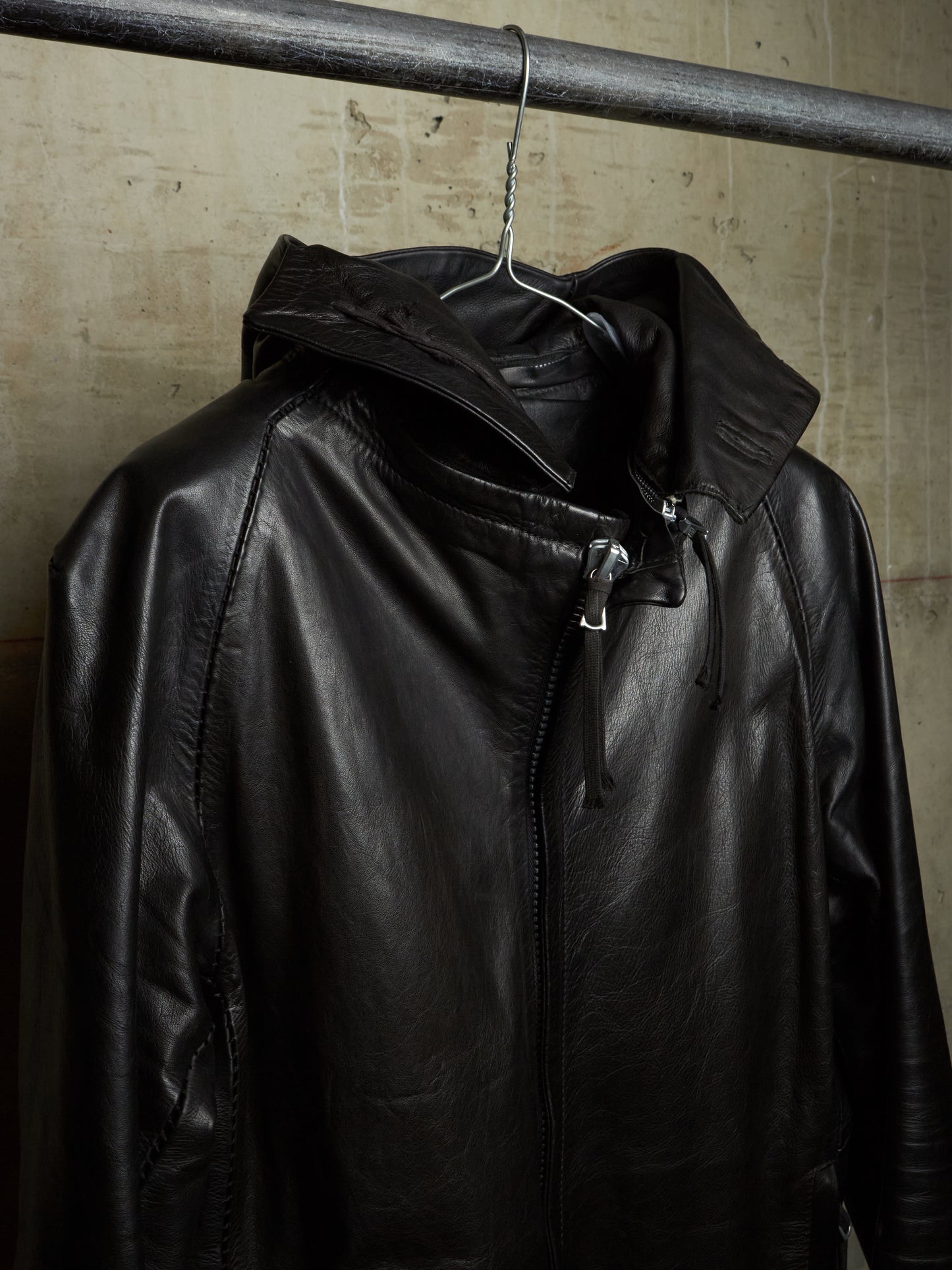A black leather hooded jacket hanging on a metal hanger from a dark industrial rod, set against a textured beige concrete wall.