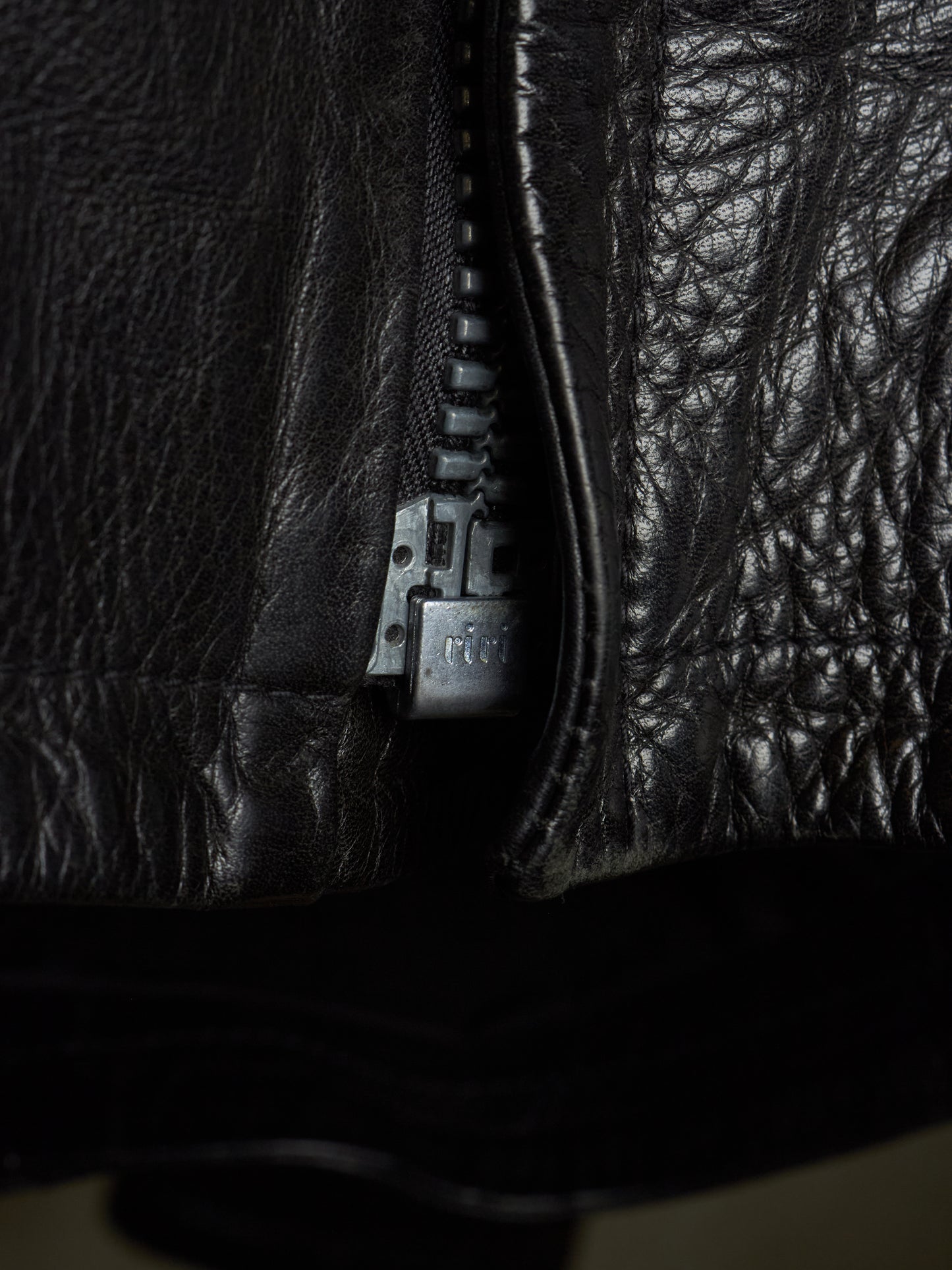 Close-up of a black leather jacket with a silver-toned metal zipper pull, showing the textured leather material in dramatic lighting.
