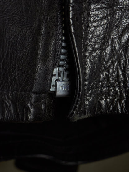 Close-up of a black leather jacket with a silver-toned metal zipper pull, showing the textured leather material in dramatic lighting.