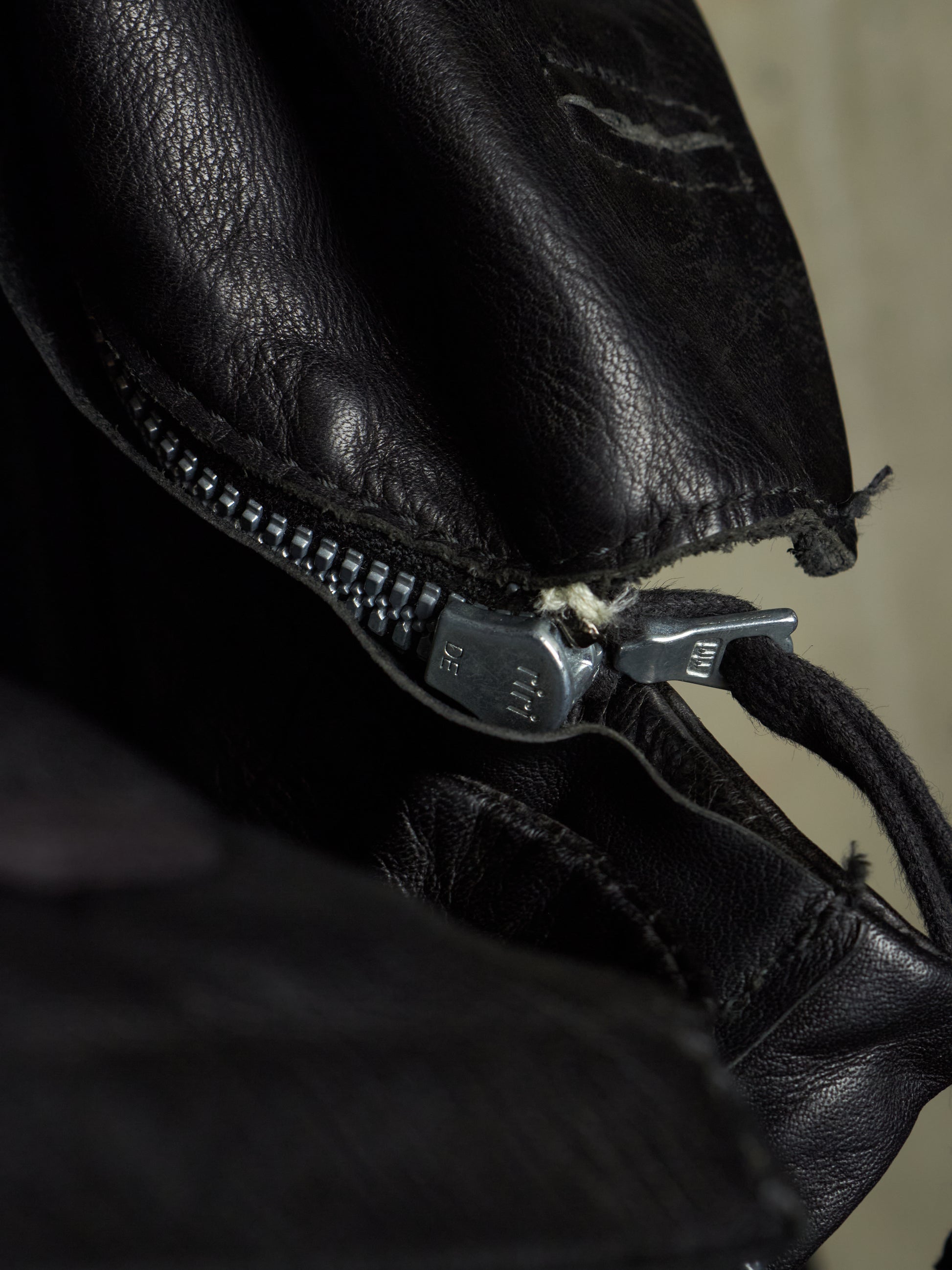 Close-up of a black leather bag with a silver metal zipper and pull tab, showing the texture of the leather and a small stitched detail.