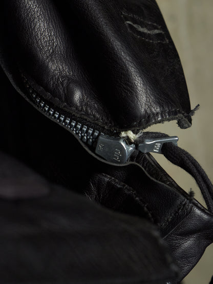 Close-up of a black leather bag with a silver metal zipper and pull tab, showing the texture of the leather and a small stitched detail.