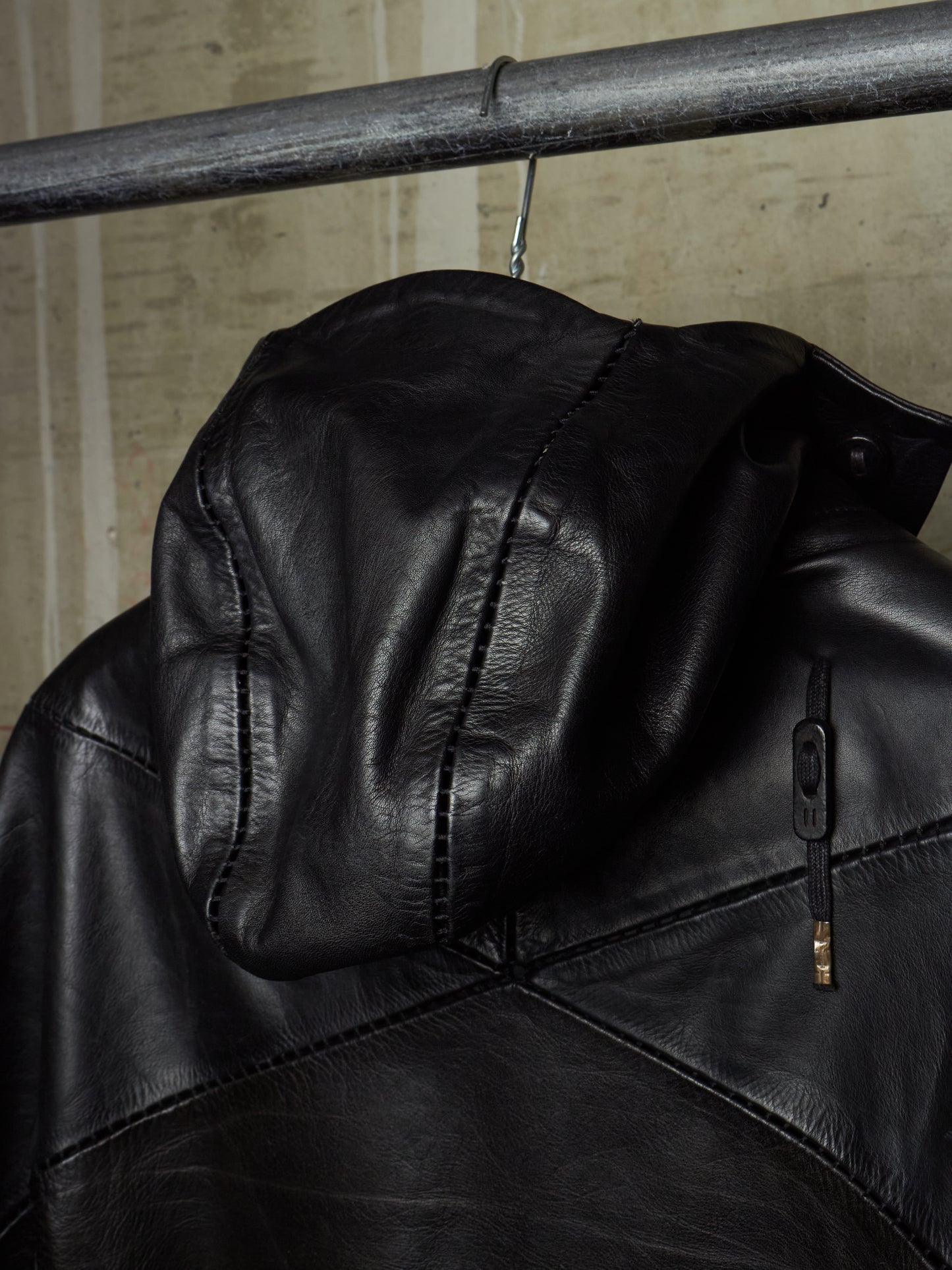 A close-up of a black leather hooded jacket hanging from a metal rod against a textured beige wall.