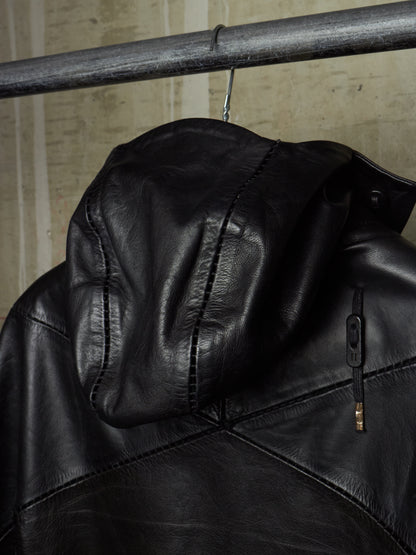A close-up of a black leather hooded jacket hanging from a metal rod against a textured beige wall.